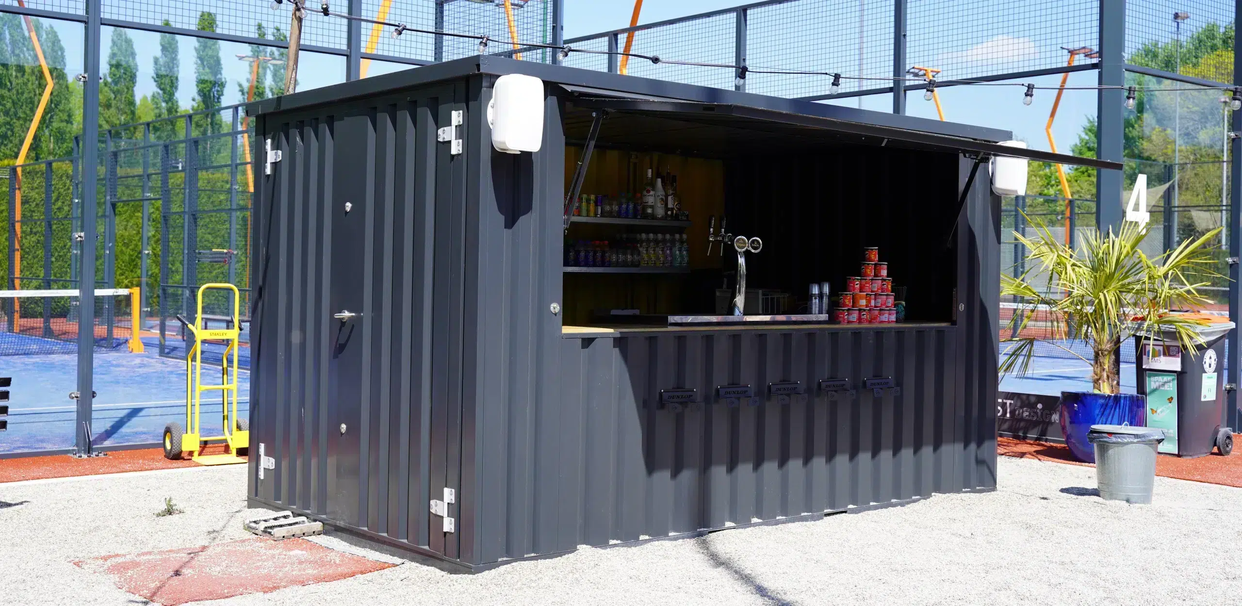 Antraciete barcontainer met een open klep naast padel velden